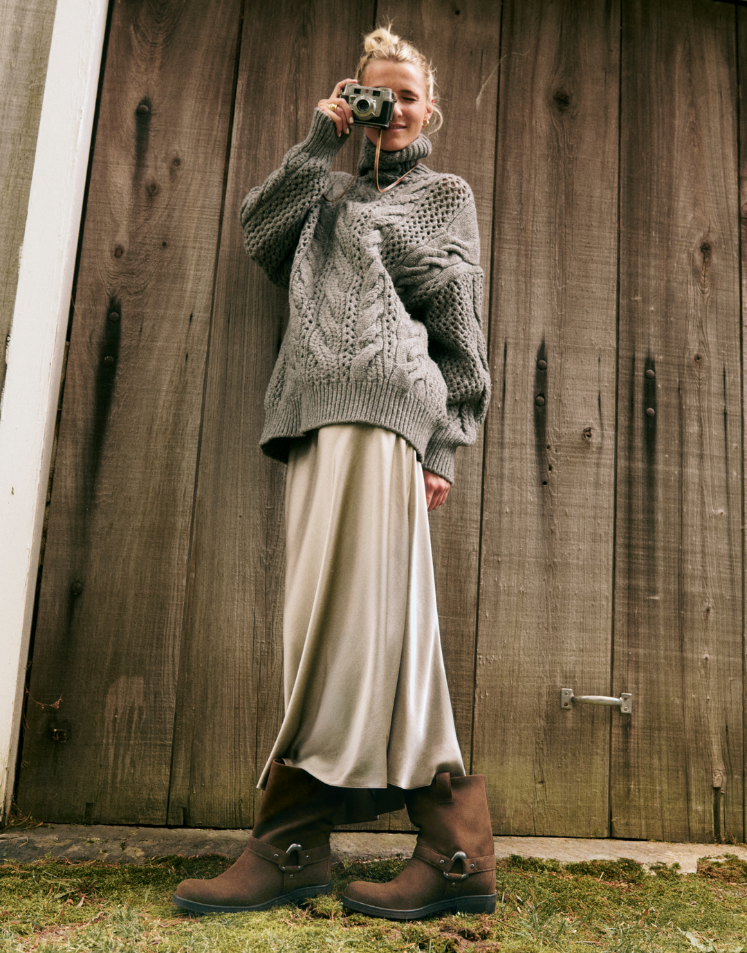 woman standing outside in front of wooden door wearing long silk skirt, cozy sweater in brown moto boots taking a picture with camera