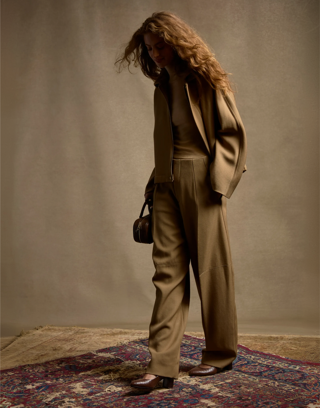 A model stands on a patterned rug against a textured beige wall, wearing a matching taupe-brown tailored suit with a blazer, wide-leg trousers, and brown leather shoes, while holding a small dark handbag.