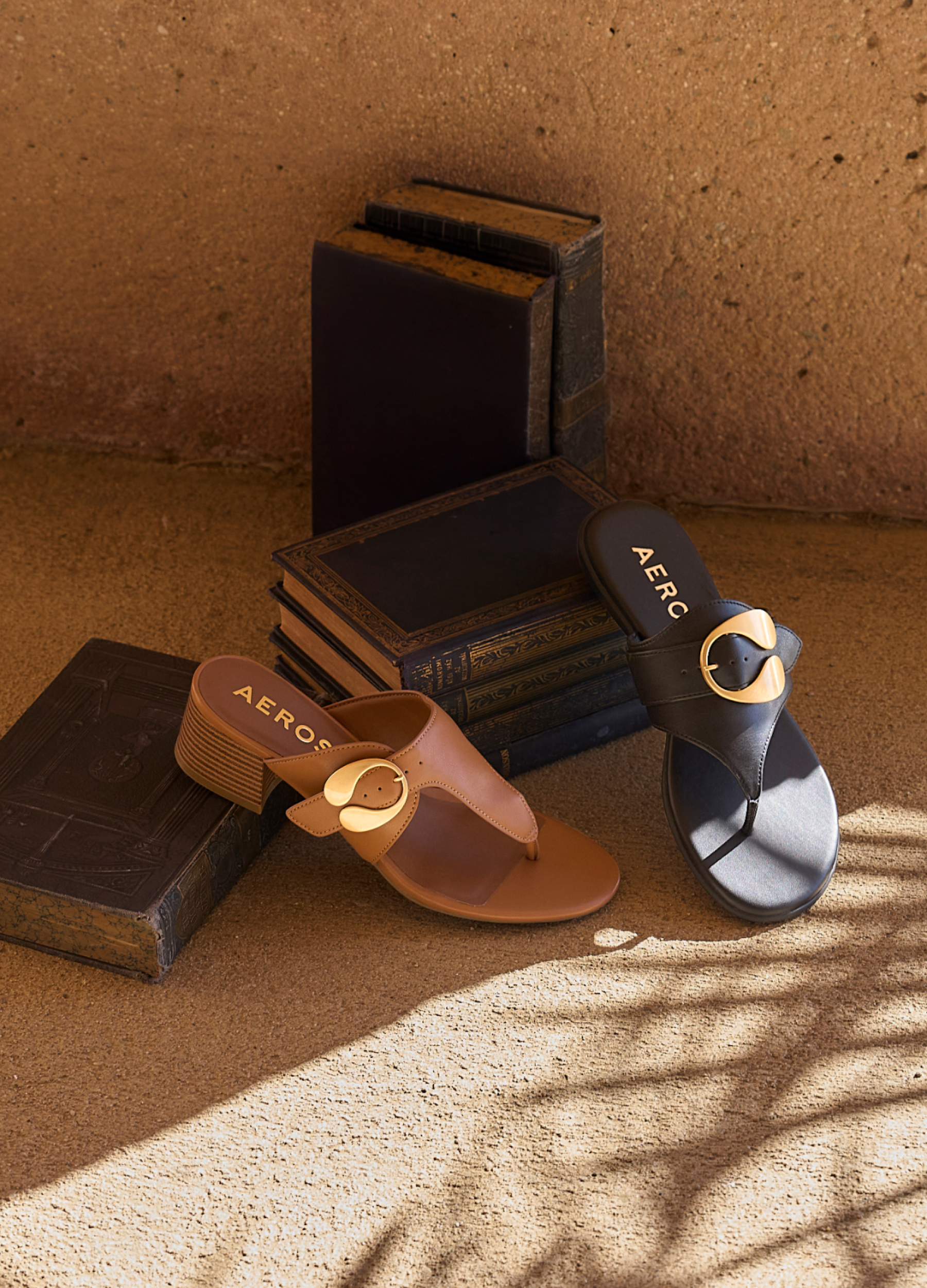 A pair of AEROSOLES brand slide sandals styled on a textured brown surface with vintage books. One sandal is tan with a gold-tone circular buckle, and the other is black with a matching black circular buckle.