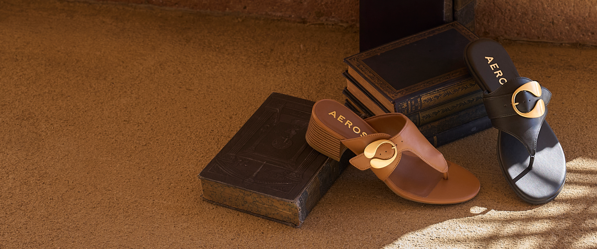 A pair of AEROSOLES brand slide sandals styled on a textured brown surface with vintage books. One sandal is tan with a gold-tone circular buckle, and the other is black with a matching black circular buckle.