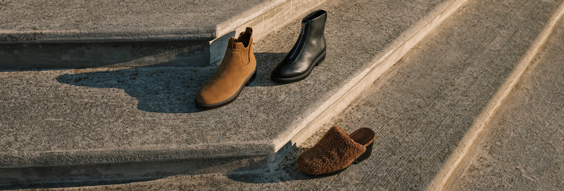 Two pair of boots and one heeled mule on a concrete surface with a step.