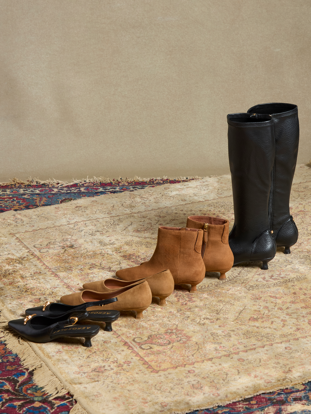 A styled collection of women's footwear arranged on a patterned rug against a neutral beige wall. The set includes black knee-high leather boots, black low-heeled mules, tan ankle boots, and tan mid-calf boots.