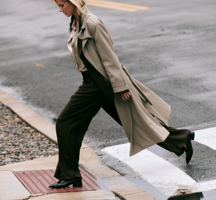 A woman walking across a city crosswalk wearing a long beige trench coat over dark brown wide-leg trousers and black shoes.
