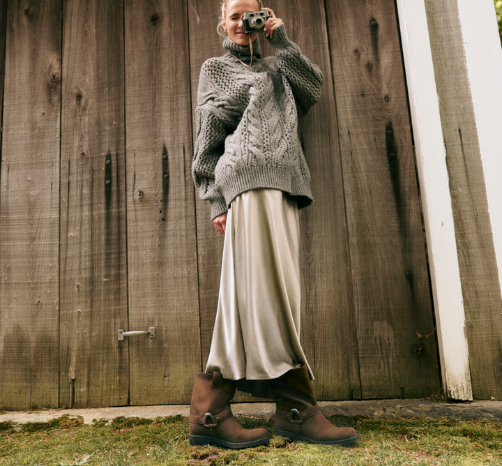 A woman stands in front of a rustic wooden door wearing a chunky gray cable-knit sweater, a cream satin skirt, and brown leather boots with buckle straps, holding a vintage camera to her face.