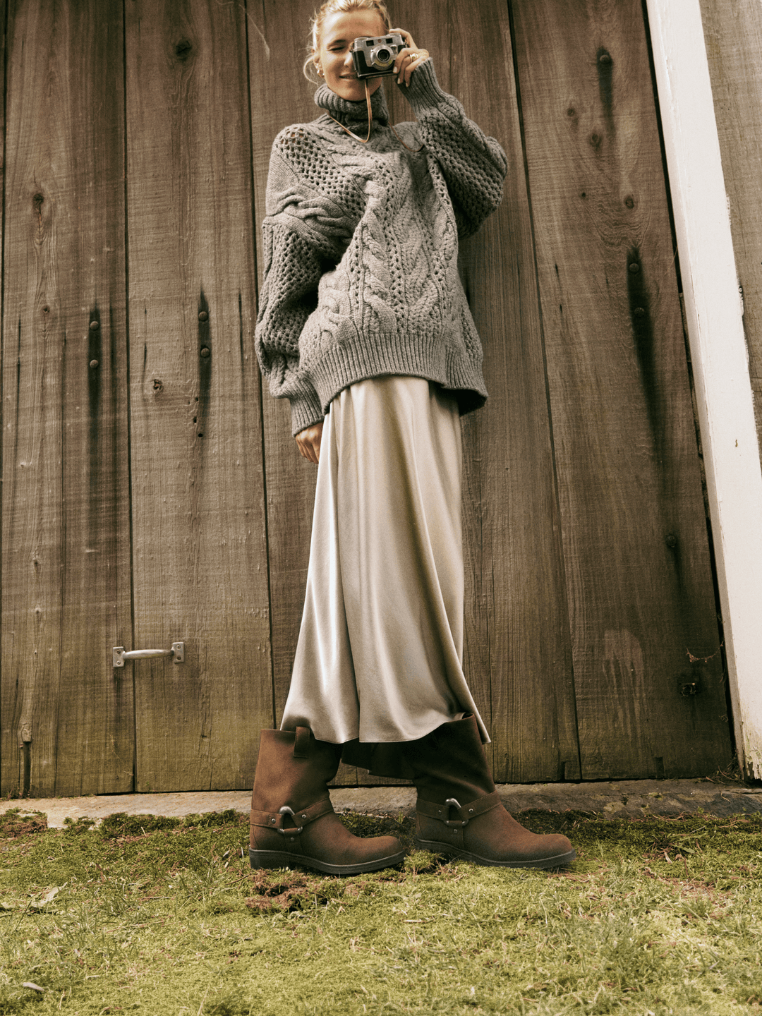 A woman stands outdoors in front of a rustic wooden door, wearing a chunky gray cable-knit turtleneck sweater, a long cream satin skirt, and brown leather boots. She is holding a vintage camera to her face.