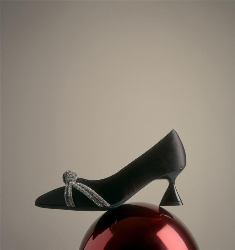 Black high-heeled shoe with a decorative bow on a reflective red surface against a beige background. the holiday shop. 