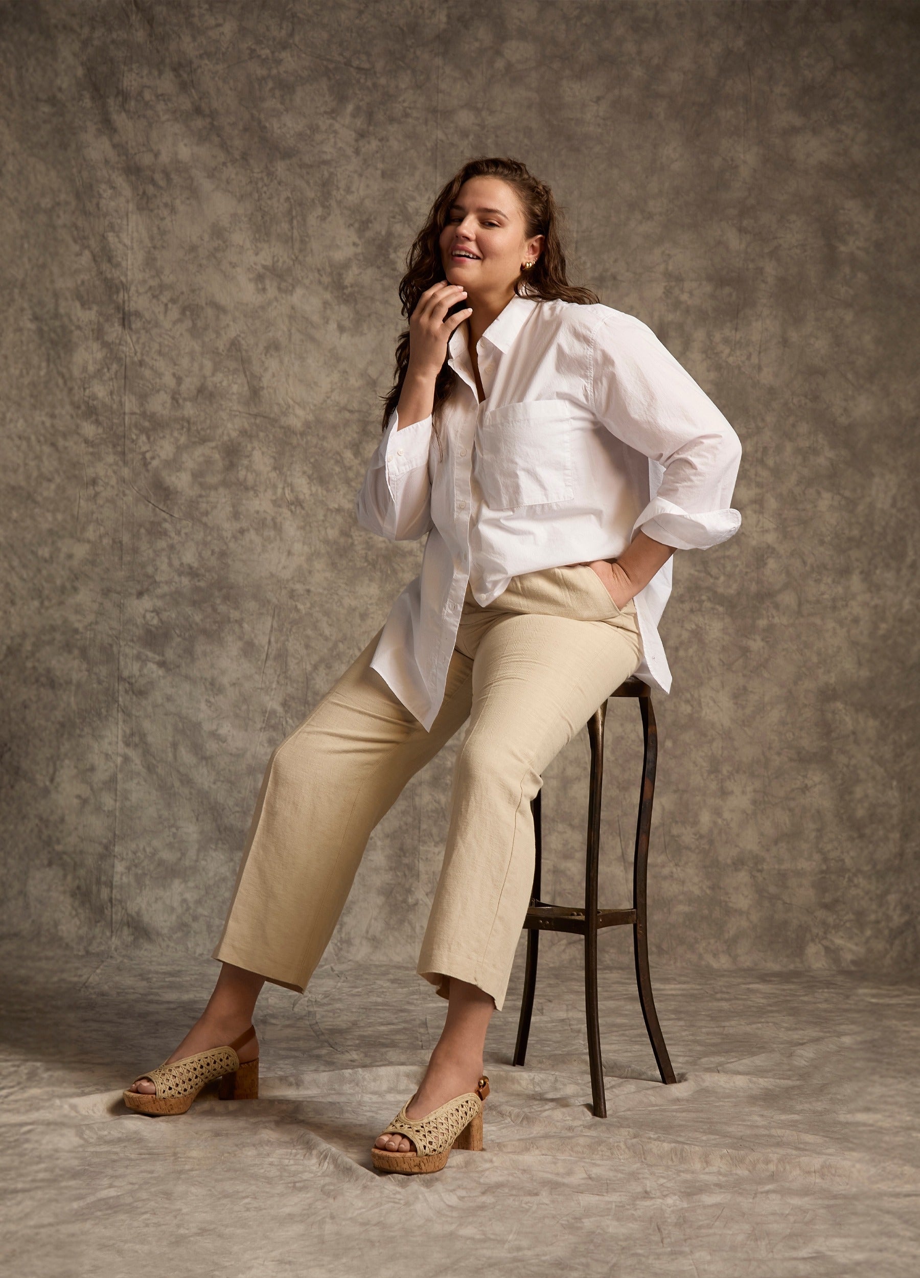 Woman sitting on a stool against a dark gray background wearing raffia peep toe heeled sandals