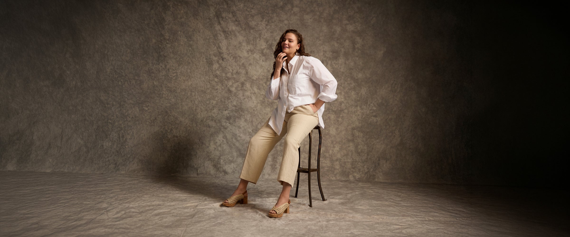 Woman sitting on a stool against a dark gray background wearing raffia peep toe heeled sandals