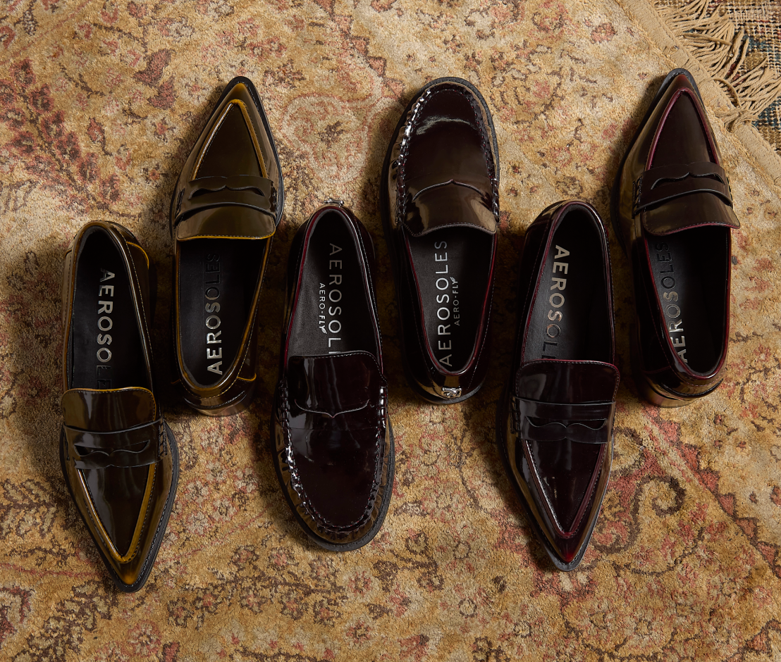Six leather penny loafers in various dark shades, arranged in a fan-like pattern on a vintage patterned rug. The shoes feature glossy patent leather finishes in black, dark brown, and deep burgundy, with contrasting stitching and interior branding.
