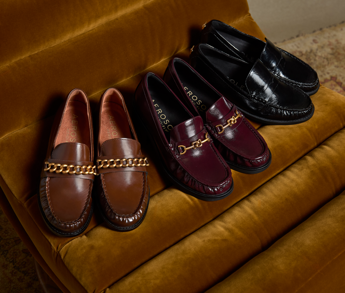 Four loafers from the brand CROSS, arranged on a plush mustard velvet chair. The shoes come in brown, burgundy, and black leather, each featuring a decorative gold chain across the vamp.