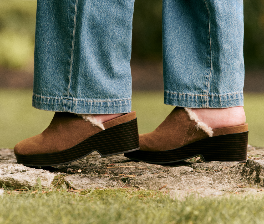 A close-up of a person wearing brown suede clogs with a soft cream shearling lining, paired with blue jeans, standing on a stone outdoors.