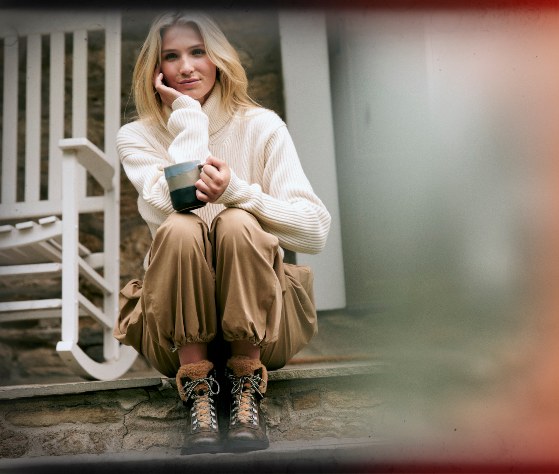 A woman sits on stone steps wearing a cream ribbed turtleneck sweater, tan cargo pants, and brown lace-up boots, holding a dark mug. A white rocking chair is visible in the background.