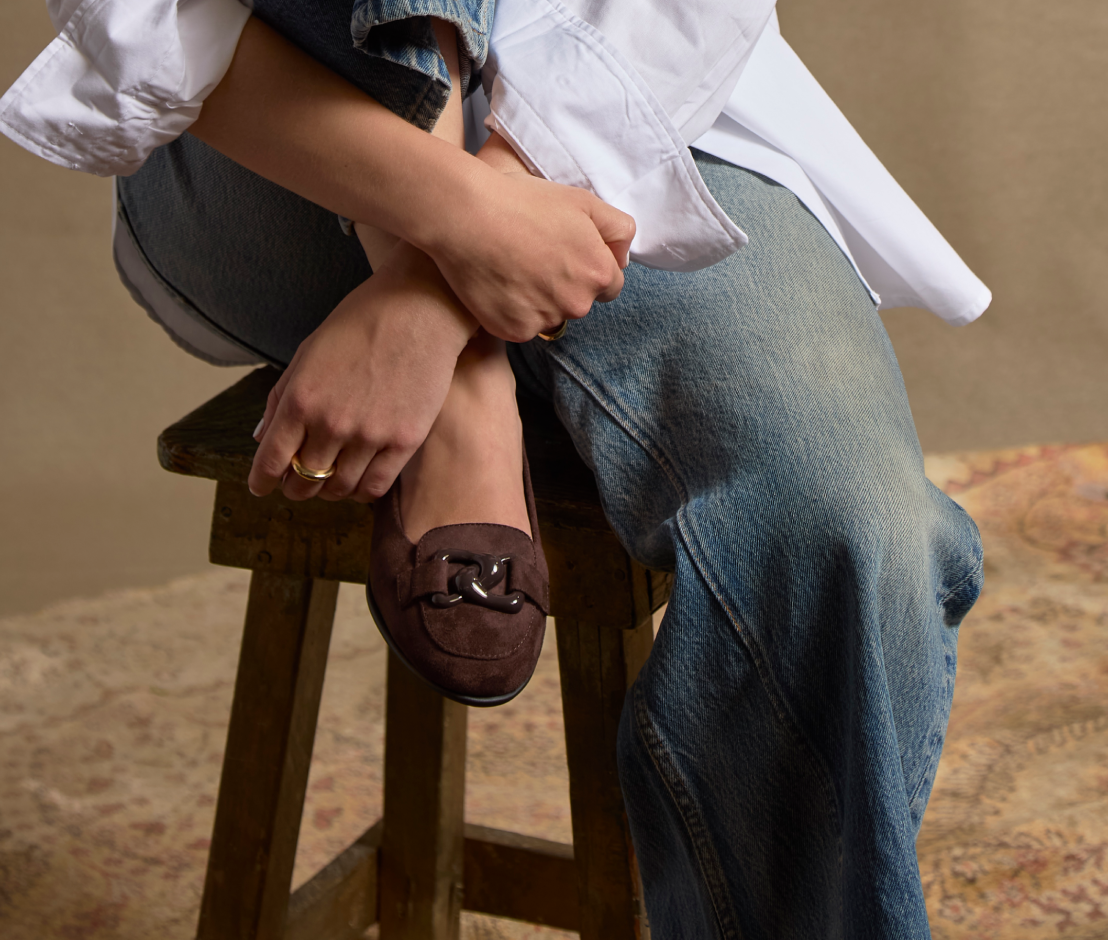 A person seated on a wooden stool wearing a dark brown suede loafer with a decorative front ornament, blue jeans, and a white shirt, against a neutral beige background.