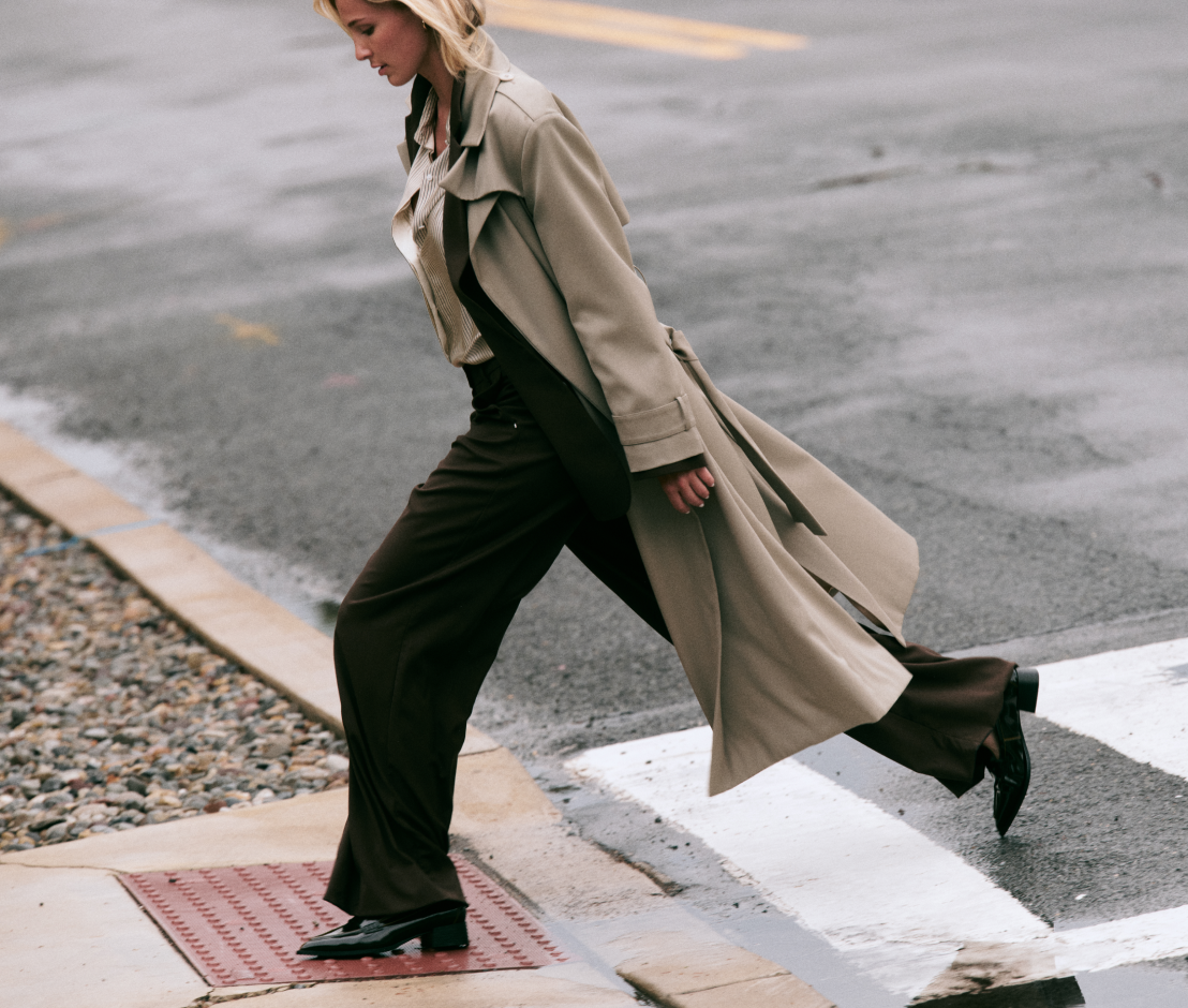 A woman walking across a crosswalk on a wet city street, wearing a long beige trench coat over a dark brown outfit and black shoes.