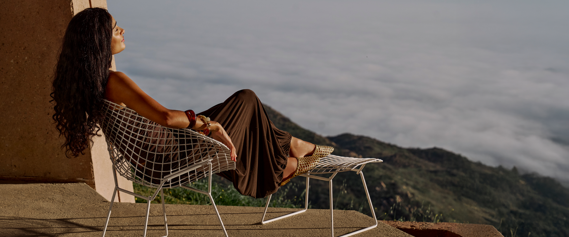 Made to Get Away. The newest arrivals, made for exploring in style. Woman sitting on a metal chair with a scenic view of mountains and clouds.