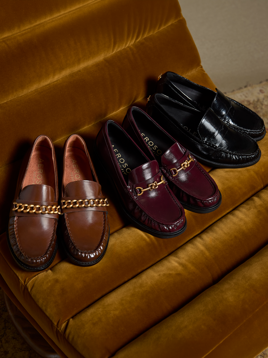 Four loafers arranged on a mustard velvet chair: two brown with a gold chain strap, two burgundy with a gold chain strap, and two black. The burgundy pair shows the brand name EROS.