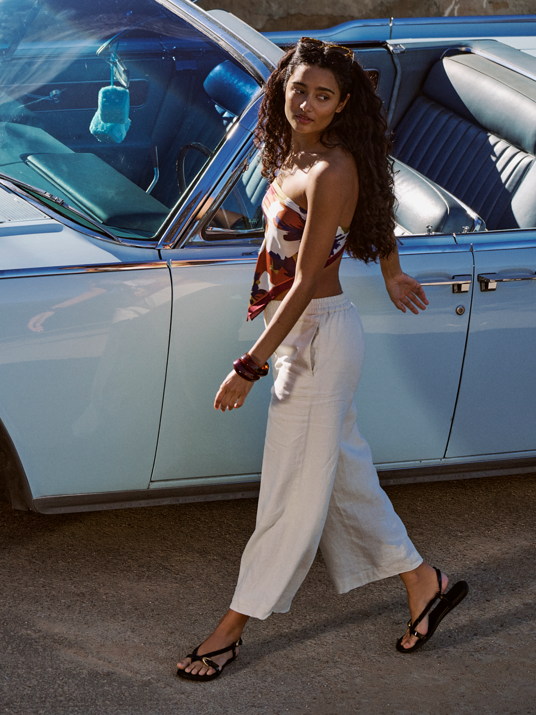TRAVEL BOUND. Effortless styles to throw on and go. Woman standing next to a vintage car, wearing a patterned top and white pants.