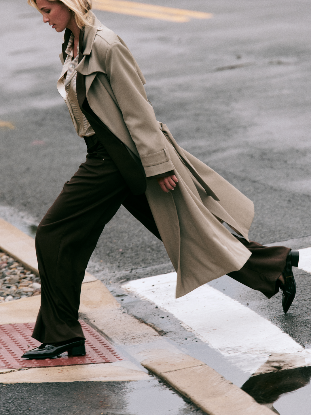 A woman walking across a wet city crosswalk wearing a long beige trench coat over dark brown wide-leg trousers and black loafers.