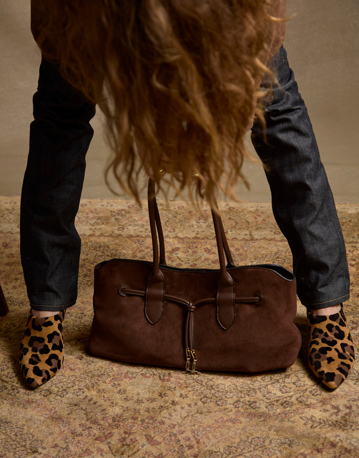 A brown suede shoulder bag with dark brown leather straps and a gold-tone zipper, placed on a patterned rug between a person's legs. The person is wearing dark blue jeans and leopard-print pointed shoes.