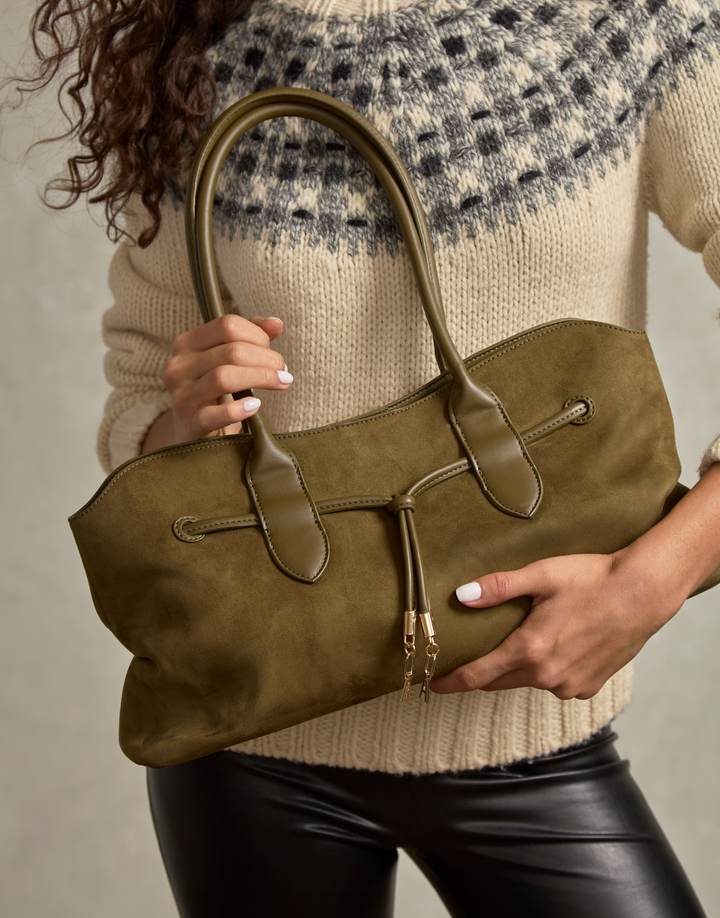 A woman holds a large olive green suede tote bag with top handles and a front flap detail, wearing a cream knit sweater and black pants against a neutral studio background.