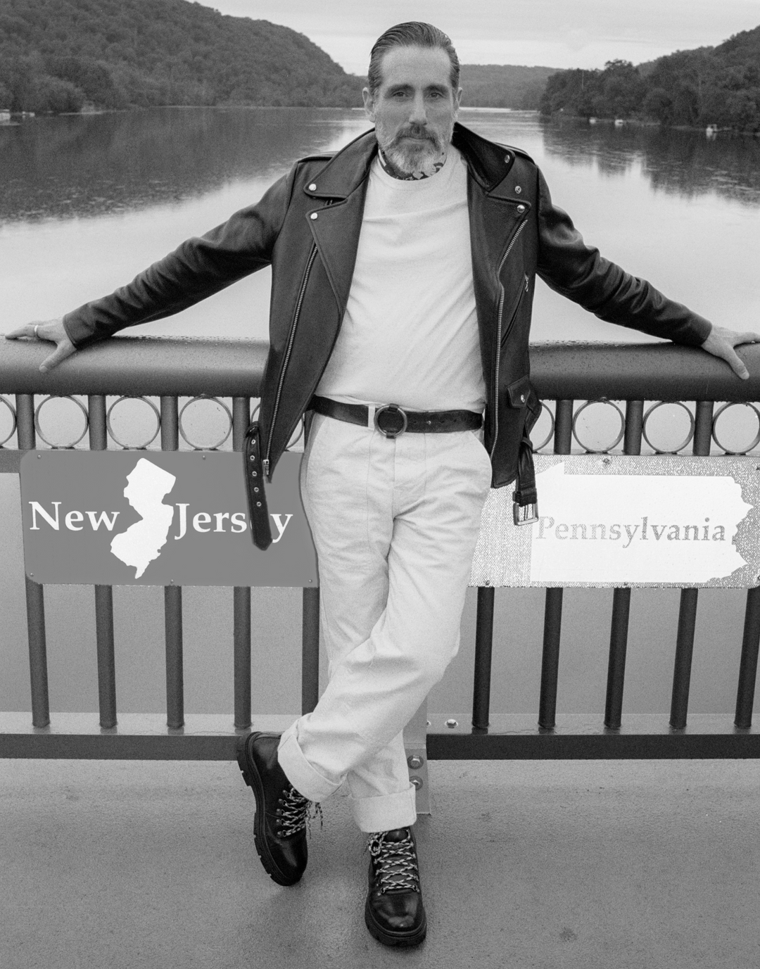 A man wearing a black leather moto jacket, white t-shirt, white pants, and black boots leans against a bridge railing with a lake and forest in the background. Two graphic overlays show the states of New Jersey and Pennsylvania.