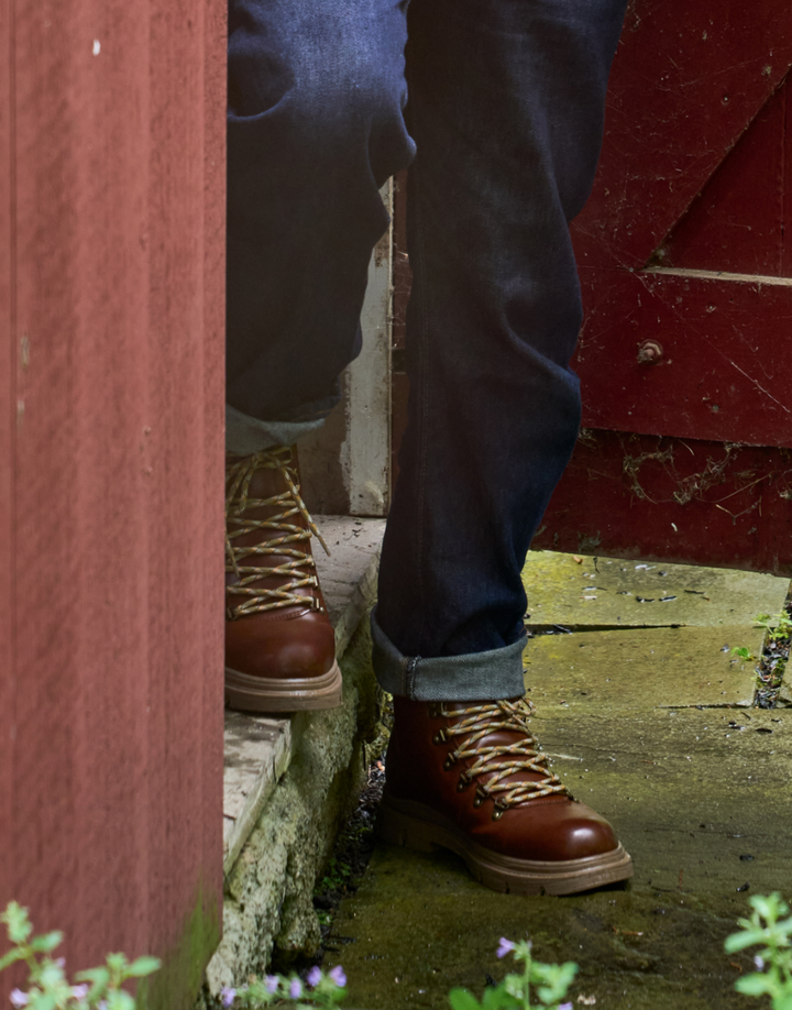 A close-up lifestyle shot of a person wearing brown leather lace-up work boots with tan laces and dark blue jeans, stepping onto a stone step in an outdoor rustic setting.