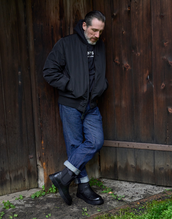 A man wearing a black bomber jacket, black t-shirt, blue jeans, and black Chelsea boots leans against a dark wooden door.