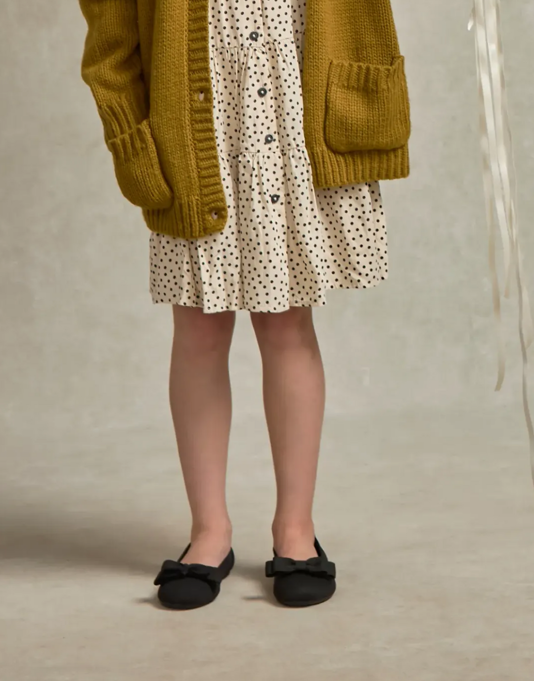 A child wearing a mustard yellow knit cardigan, a cream dress with black polka dots, and black bow flats, standing against a neutral textured beige background.