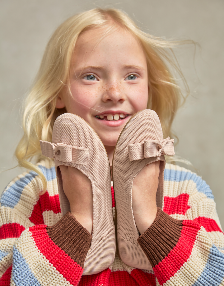 A young blonde girl with freckles smiles while holding a pair of beige ballet flats in front of her face. She is wearing a colorful striped knit sweater against a soft neutral studio background.