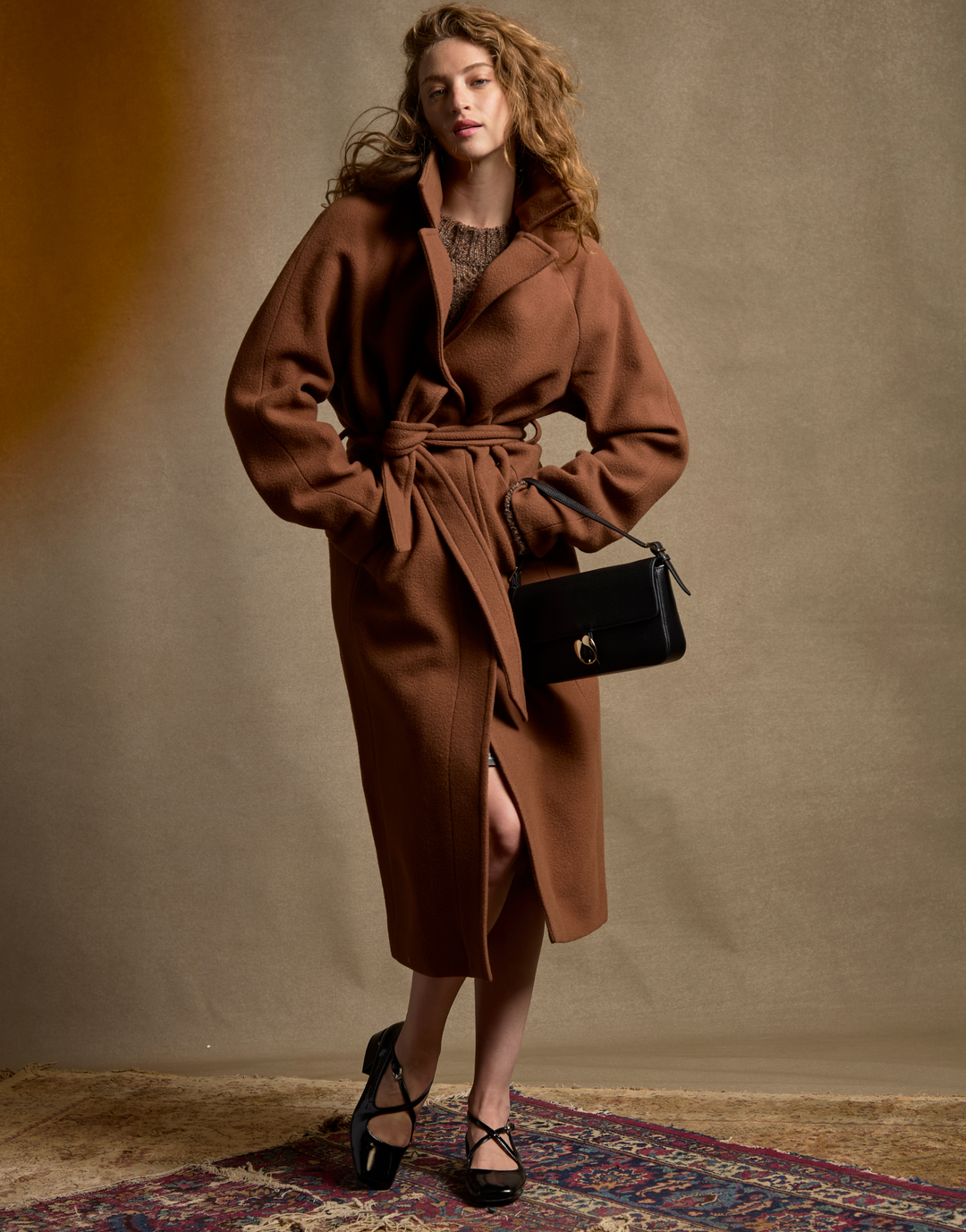 A woman models a long brown wool coat with a matching waist tie, styled with a black leather handbag and black strappy heels against a textured beige studio backdrop.