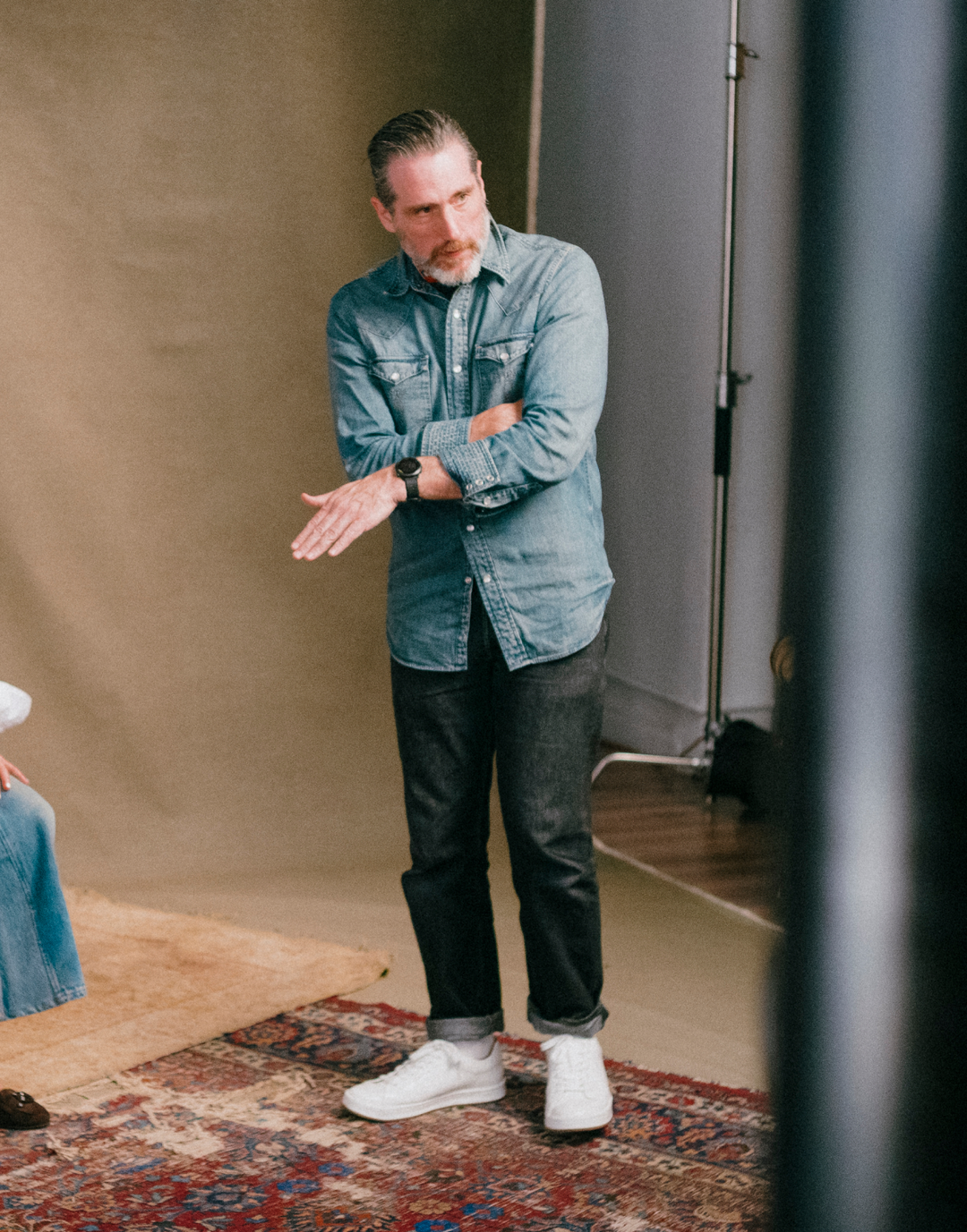 A man wearing a light blue denim button-up shirt, dark gray jeans, and white sneakers stands in a studio setting with a beige backdrop and a patterned rug.