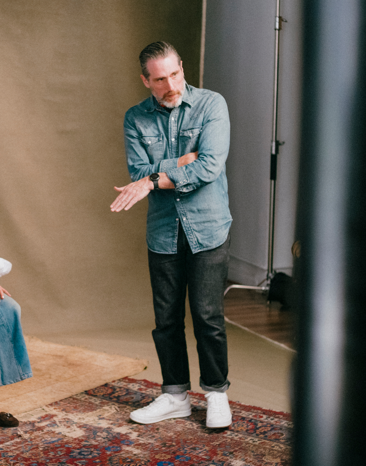 A man wearing a light blue denim button-up shirt, dark gray jeans, and white sneakers stands in a studio setting with a beige backdrop and a patterned rug.