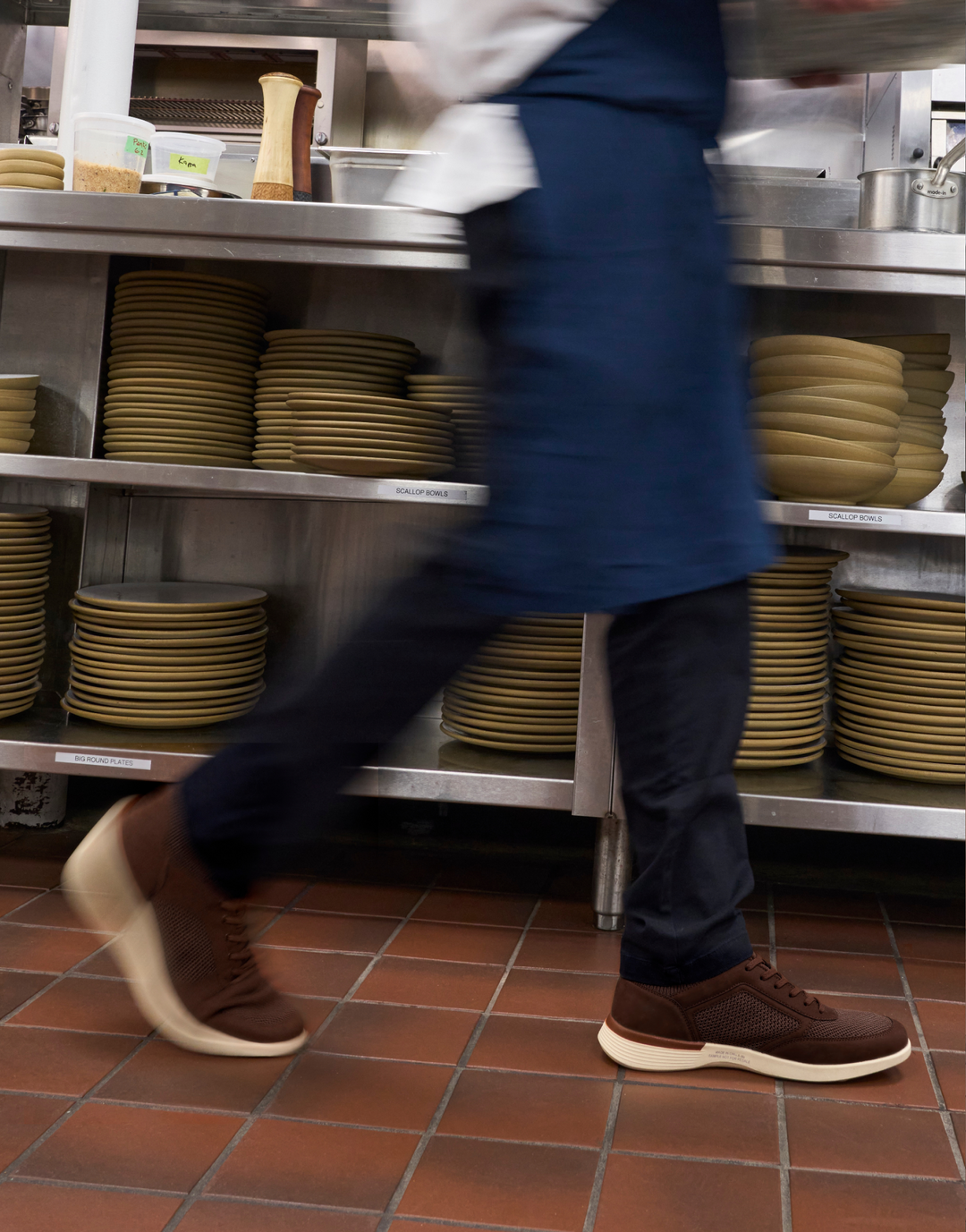 A person in a navy apron and dark pants walks across a commercial kitchen floor, with stacks of beige plates on stainless steel shelves in the background.