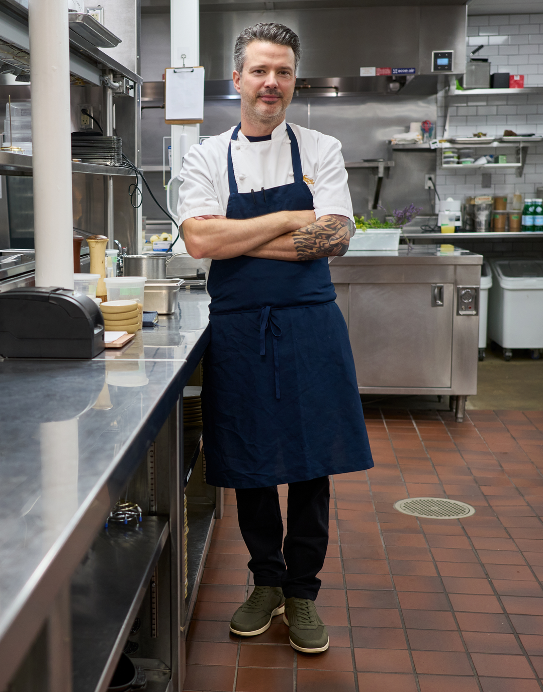 A man wearing a white chef's coat and a navy blue apron stands with arms crossed in a commercial kitchen.