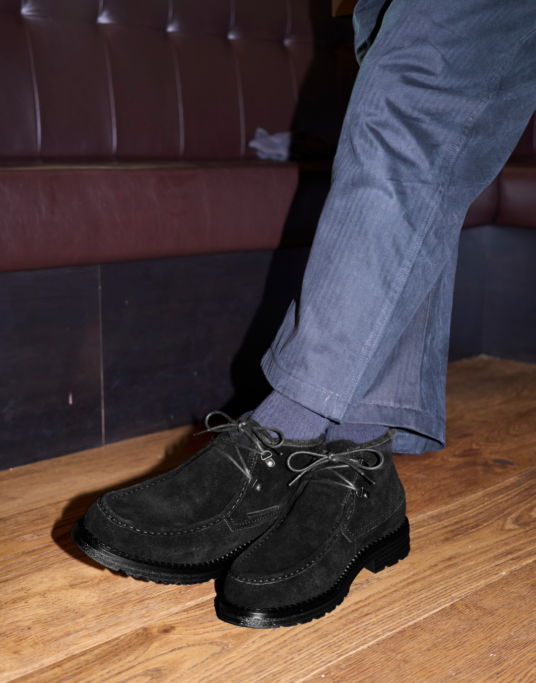 A person wearing black suede lace-up boots with gray pants, standing on a wooden floor in front of a dark leather booth.