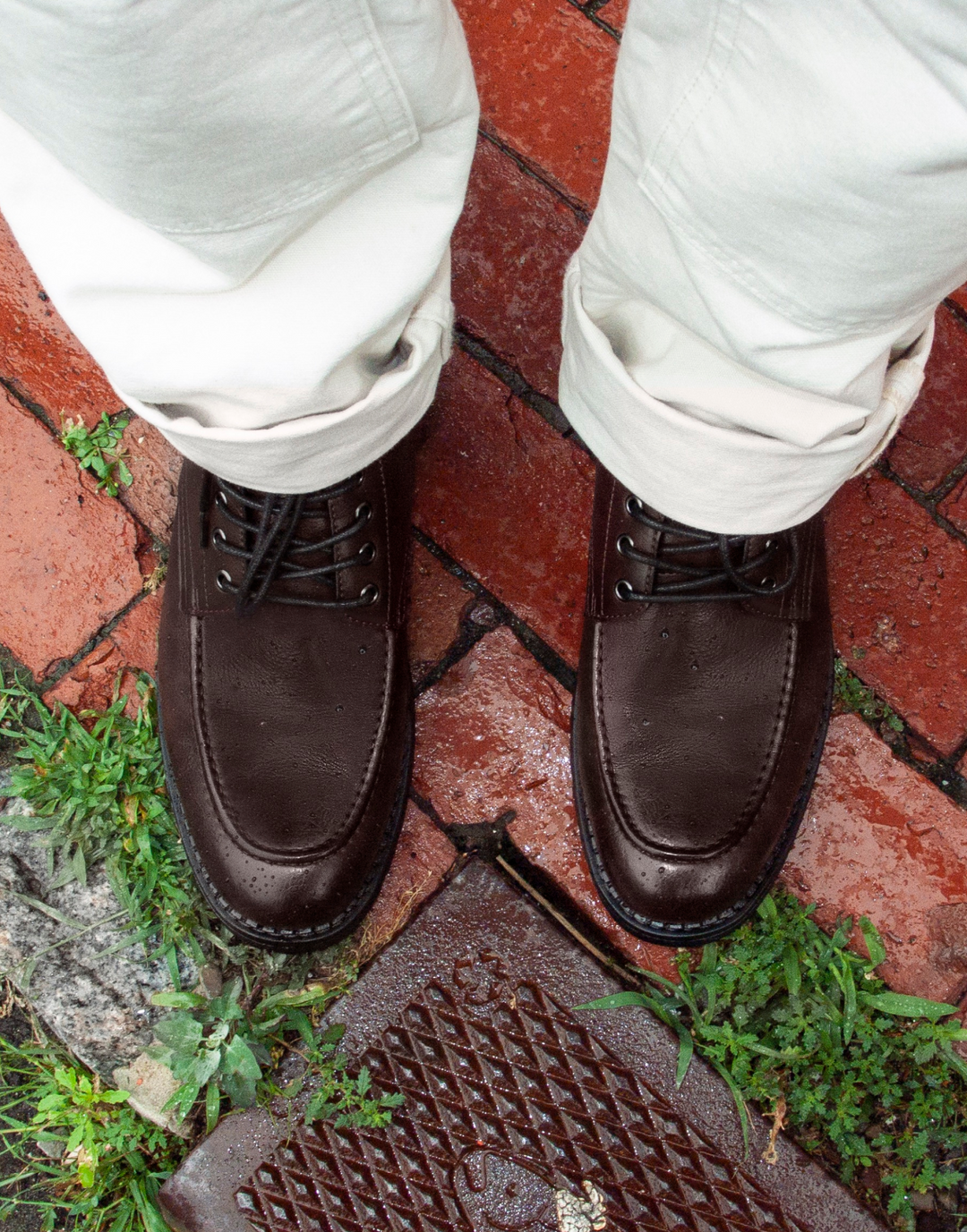 A top-down view of a person wearing dark brown leather lace-up boots and off-white trousers, standing on a red brick patio.