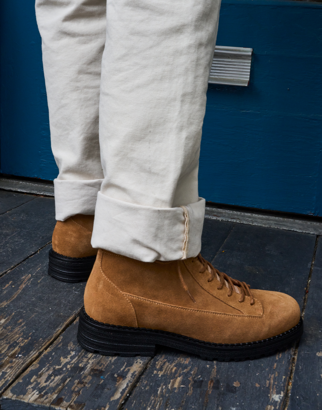 A close-up of a person's legs wearing tan suede lace-up ankle boots with black soles, paired with off-white cuffed trousers, standing on a dark wooden deck in front of a blue wall.