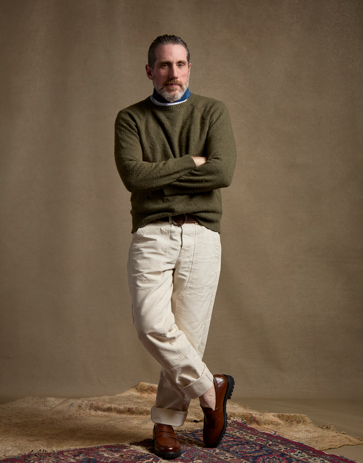 A man stands with arms crossed wearing an olive green knit sweater over a blue and white striped shirt, cream trousers, and brown leather loafers, posed against a warm brown studio backdrop with a rug at the bottom.
