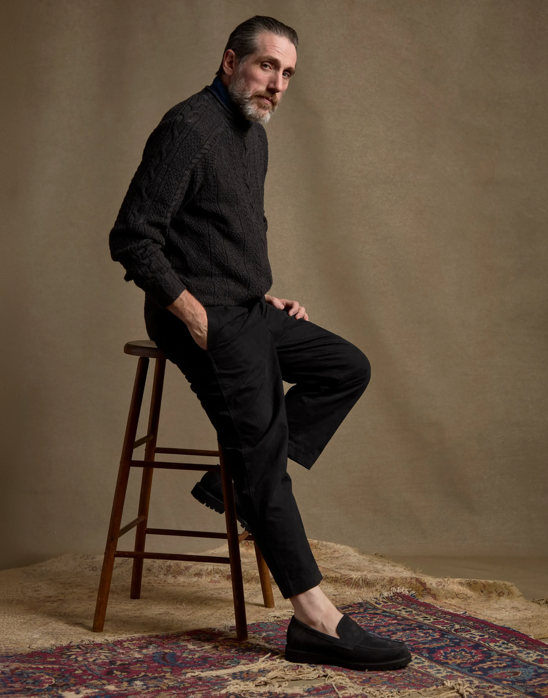 A man with a gray beard sits on a wooden stool, wearing a dark textured knit turtleneck sweater, black trousers, and black slip-on loafers against a warm brown studio backdrop.