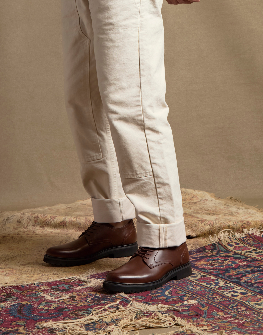A person wearing off-white trousers and dark brown leather lace-up shoes, standing on a patterned rug against a neutral beige background.