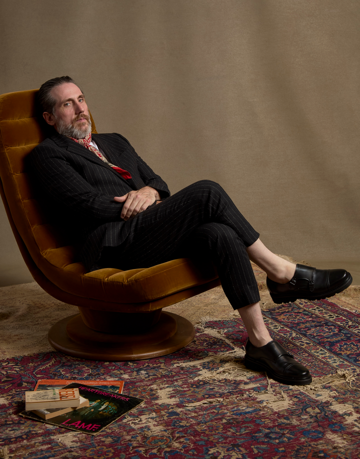 A man with a beard wearing a dark pinstripe suit, red patterned tie, and black loafers sits in a mustard yellow velvet swivel chair on a patterned rug against a beige backdrop.