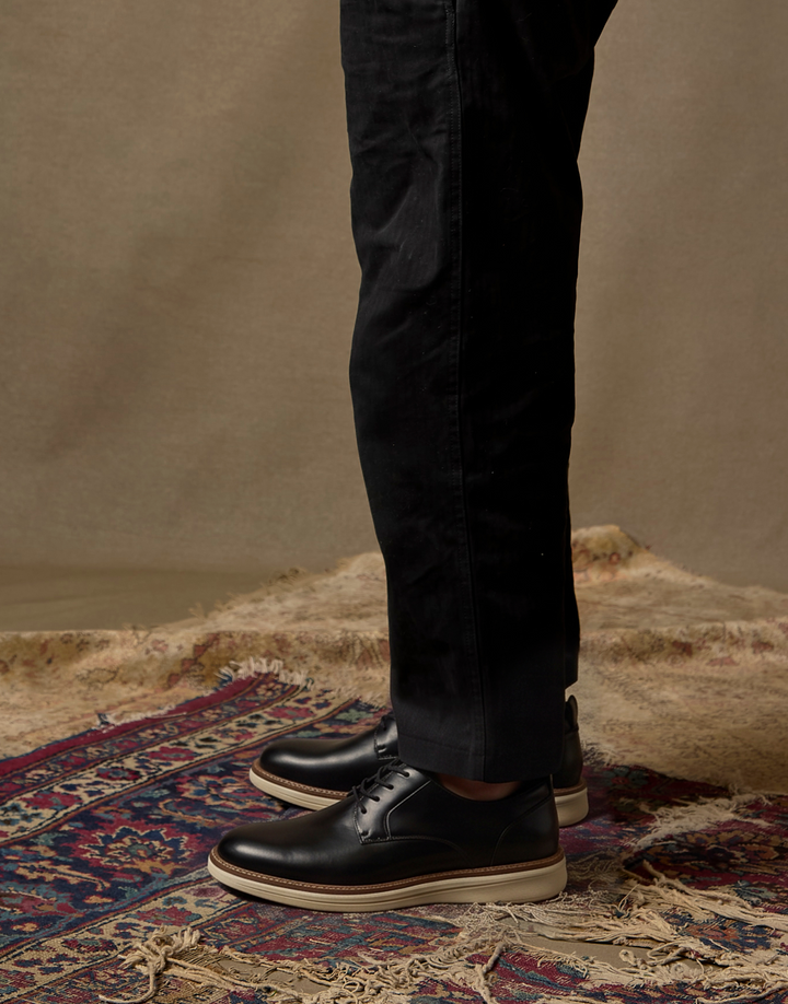 A person wearing black leather lace-up shoes with off-white soles and black trousers, standing on a patterned rug against a neutral studio backdrop.