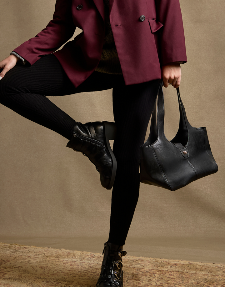A model wearing a burgundy blazer, black ribbed tights, and black ankle boots holds a large black leather shoulder bag against a textured beige backdrop.
