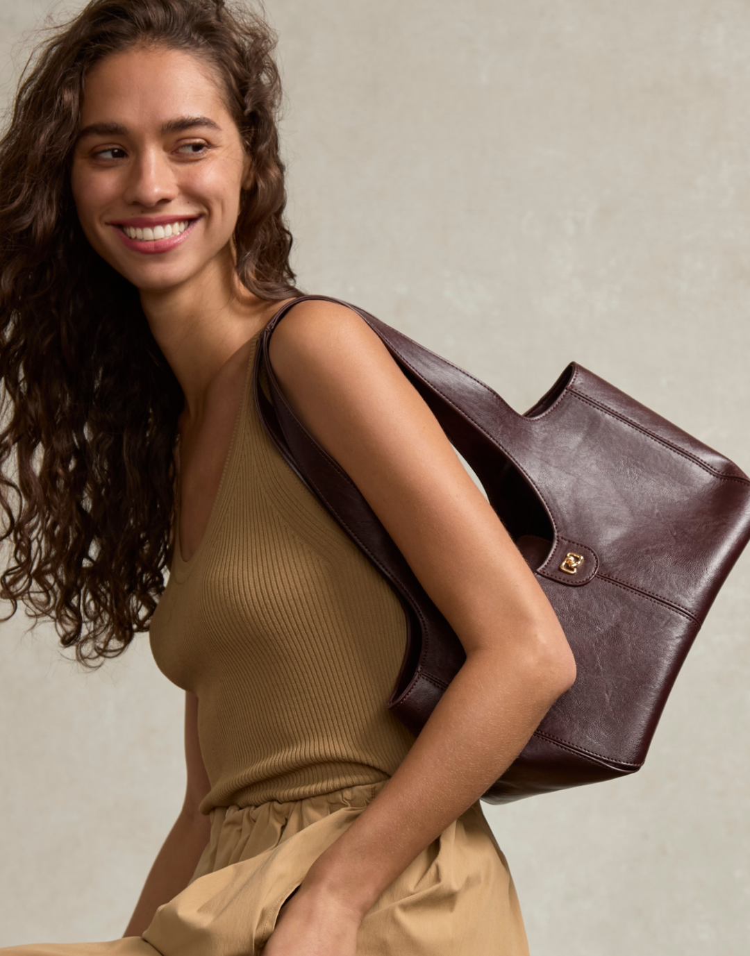A smiling woman with long curly brown hair carries a large structured tote bag over her shoulder. She is wearing a ribbed tan top and matching bottoms against a soft beige textured wall.