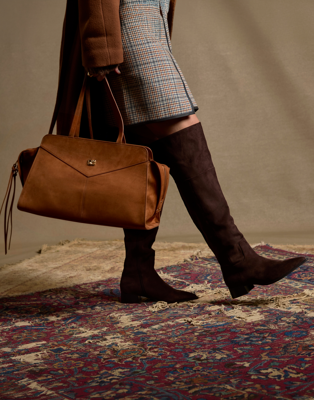 A person walking while carrying a large brown suede shoulder bag and wearing brown over-the-knee boots, with a patterned rug and neutral studio backdrop visible.
