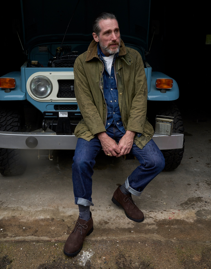 A man wearing an olive green field jacket, blue denim shirt and jeans, and brown boots sits on the front bumper of a light blue vintage off-road vehicle in a dark garage setting.