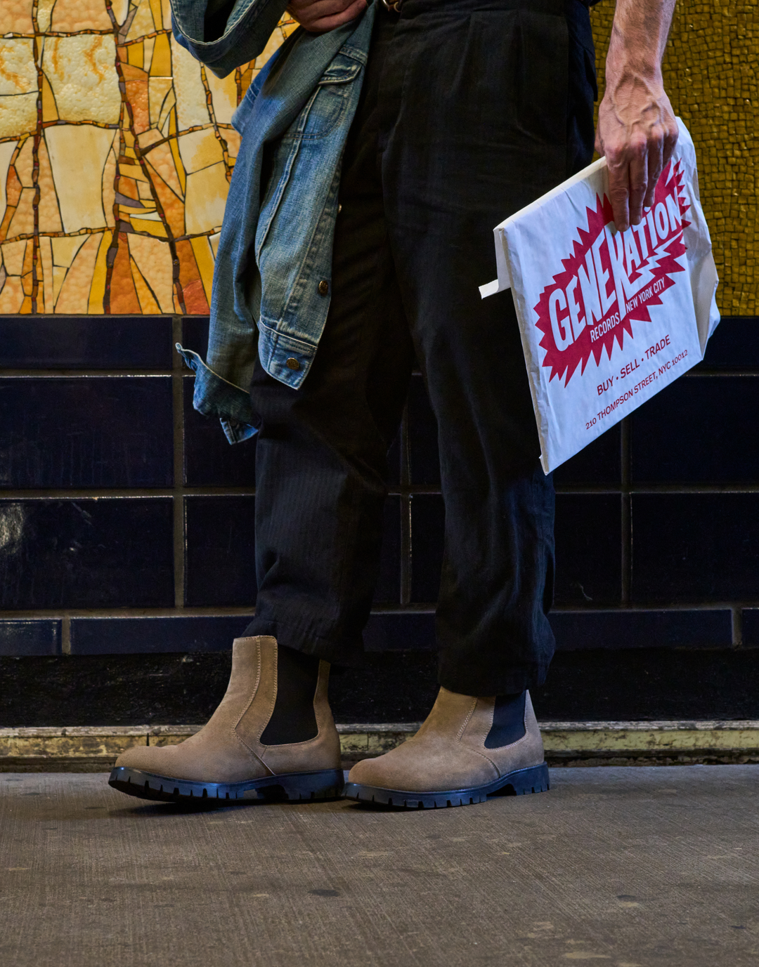 A person standing in a subway station wearing beige Chelsea-style boots, dark pants, and a faded denim jacket, holding a white Generation Boxing Club flyer.
