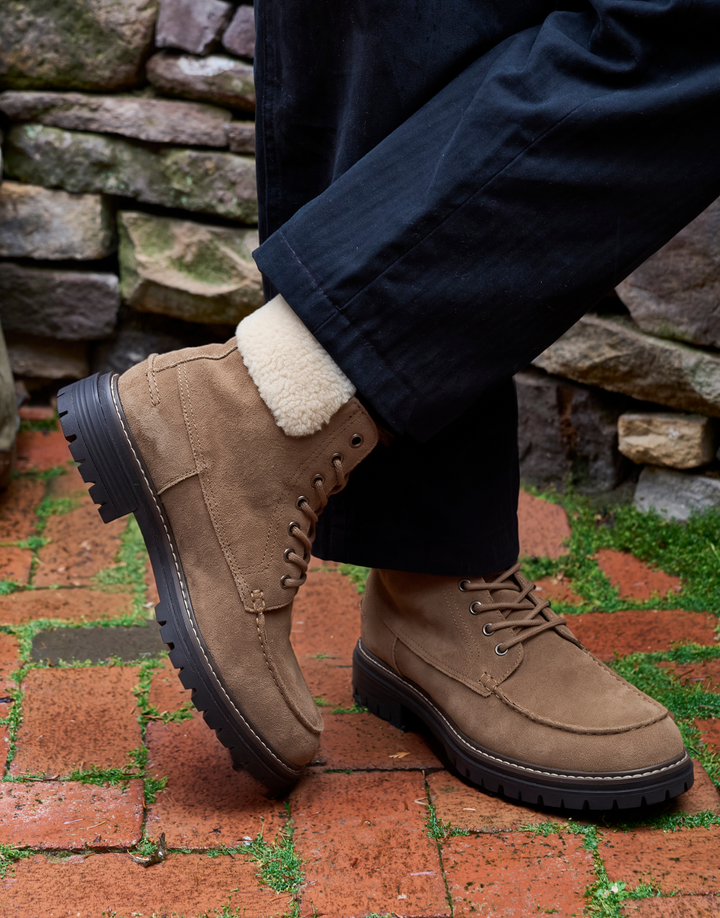 A person wearing tan suede lace-up boots with a white sherpa collar, paired with dark navy pants, standing on a red brick patio with a stone wall background.
