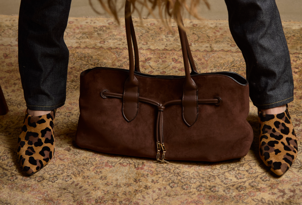 A brown suede tote bag with a drawstring closure and gold-tone zipper, shown on a patterned rug between a person's legs wearing leopard-print pointed-toe flats and dark jeans.
