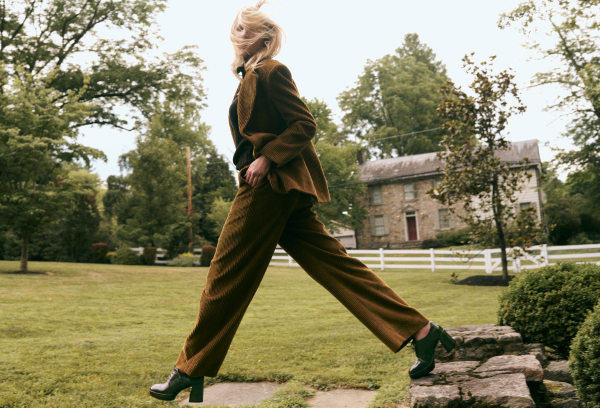 A woman wearing a brown corduroy blazer and matching wide-leg trousers walks across a grassy lawn, with trees, a stone house, and a white fence in the background.
